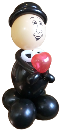 hier steht eine Ballonfigur in Form eines Bräutigam. Er hat einen schwarzen Hut, einen schwarzen Anzug, weißes Krawatte und in der Hand ein rotes Herz